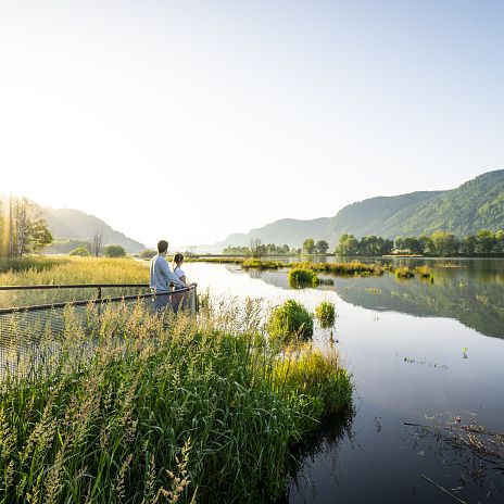 Eine Wiesen- und Teichlandschaft. Im Hintergrund sind grüne Berge zu sehen. Die Sonne scheint. Im Vordergrund eine ufernahe Aussichtsplattform zur Beobachtung. Zwei Personene stehen auf der Aussichtsplattform.